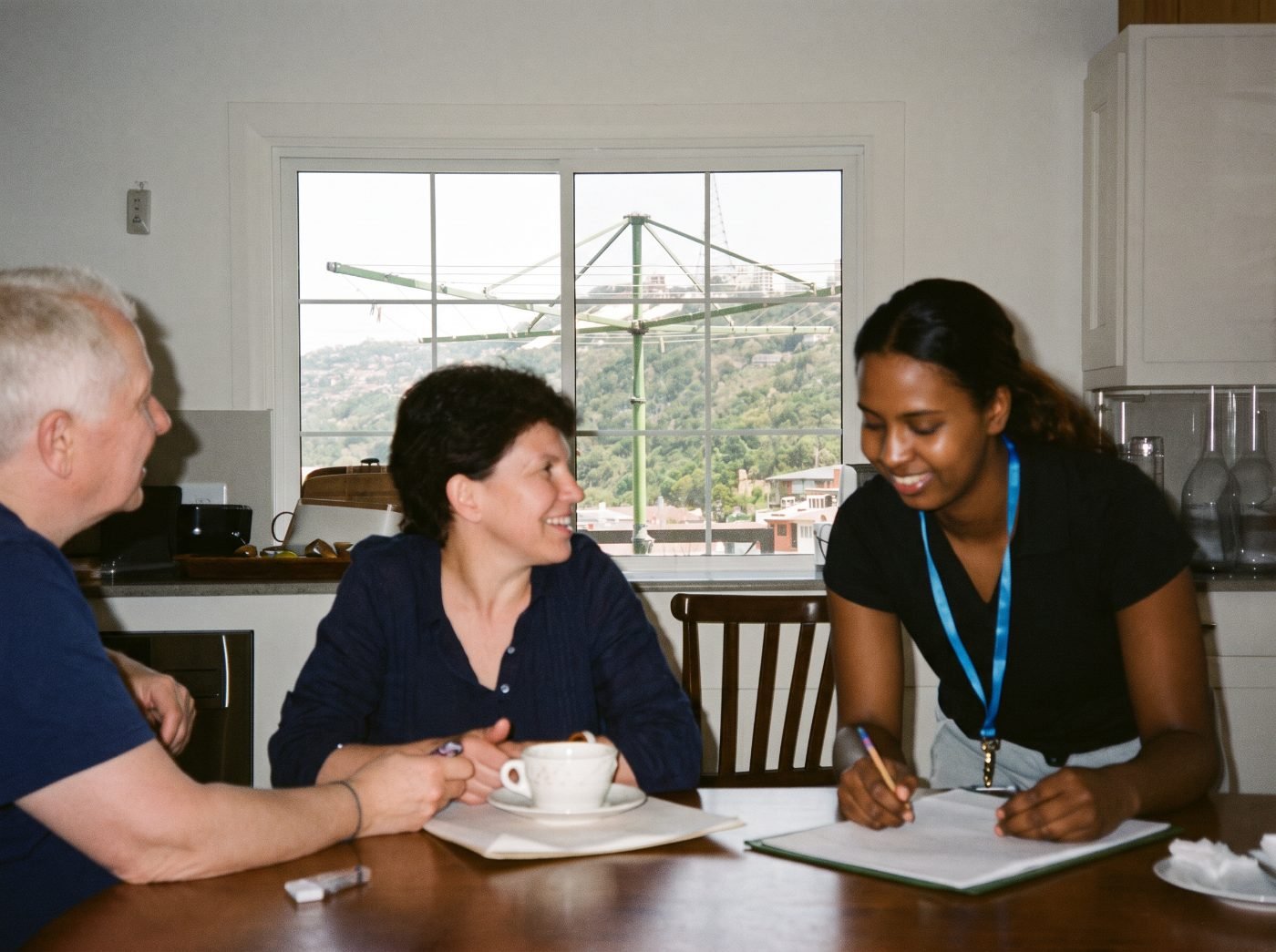 A mother, her adult daughter who has Down syndrome, and a support worker talk together at the kitchen table.