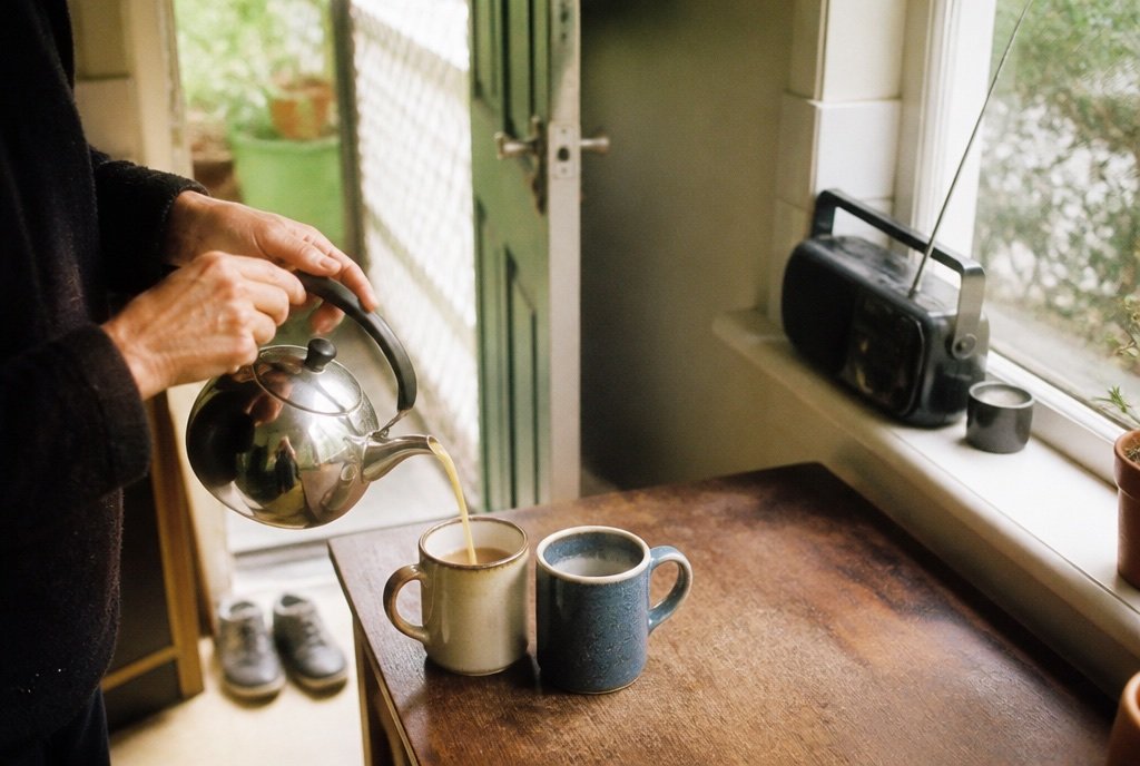 Tea being poured into two mugs on a kitchen bench, a radio on the windowsill and shoes by the door.